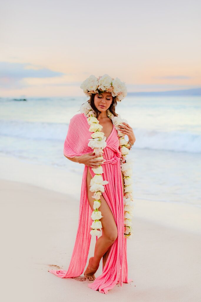 Portrait of a woman smiling softly, framed by a lush floral lei and mermaid-inspired makeup, with the early morning colors soft on the horizon.