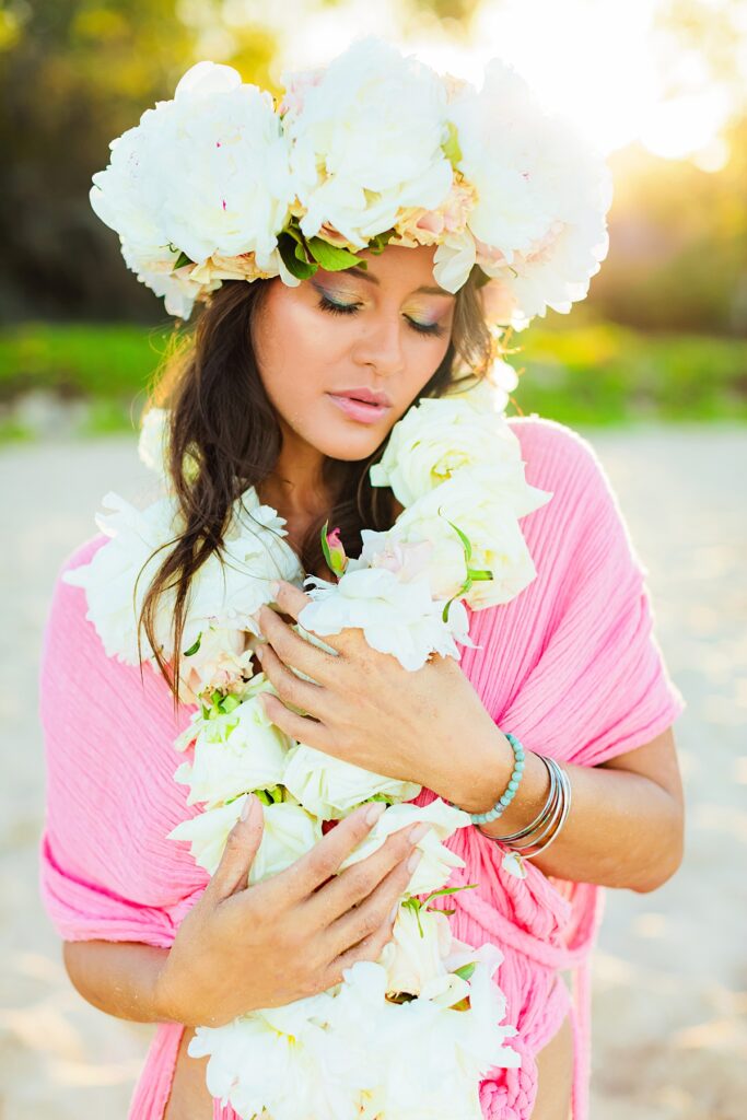 Close-up view of a woman with radiant, dewy skin, her lei po'o bursting with tropical flowers and backlit by the morning sun