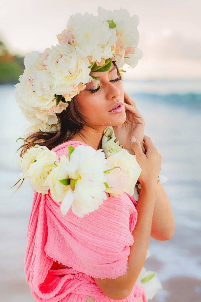 Close-up of a woman’s face, highlighting her shimmering makeup and delicate lei po’o, with the soft hues of a Maui sunrise in the background.