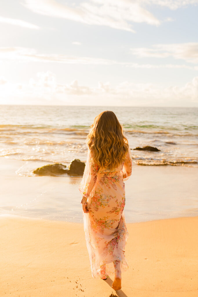Back view of a 24 weeks pregnant woman wearing a Baltic Born Layla tulle maxi dress, walking along the beach at sunset on Maui.