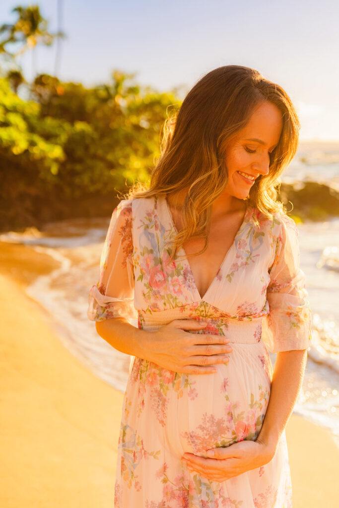 woman in flowy maternity beach photoshoot gown looks at sand while being photographed in Wailea by love and water on Maui