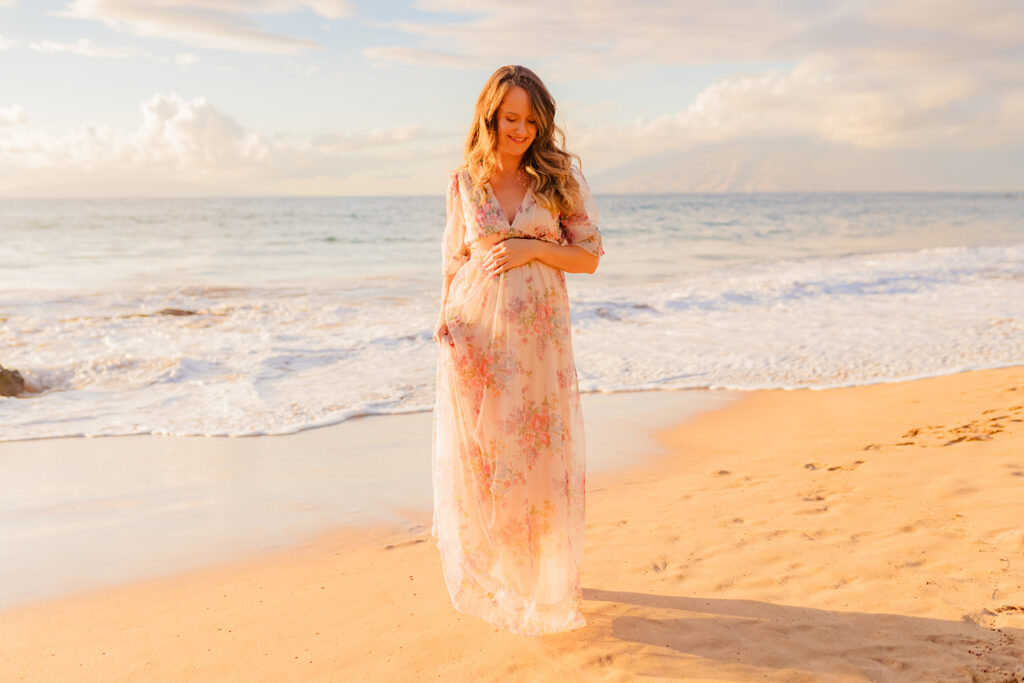 Gentle waves approach the feet of a 24 weeks pregnant woman in a flowing Baltic Born maxi dress during a maternity session on Maui.
