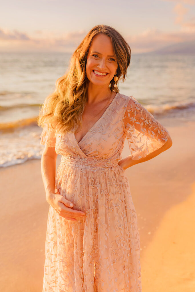 Woman holds hip and pregnant bellt while smiling at camera wearing the marseille embossed maxi dress during her beach photoshoot with love and water