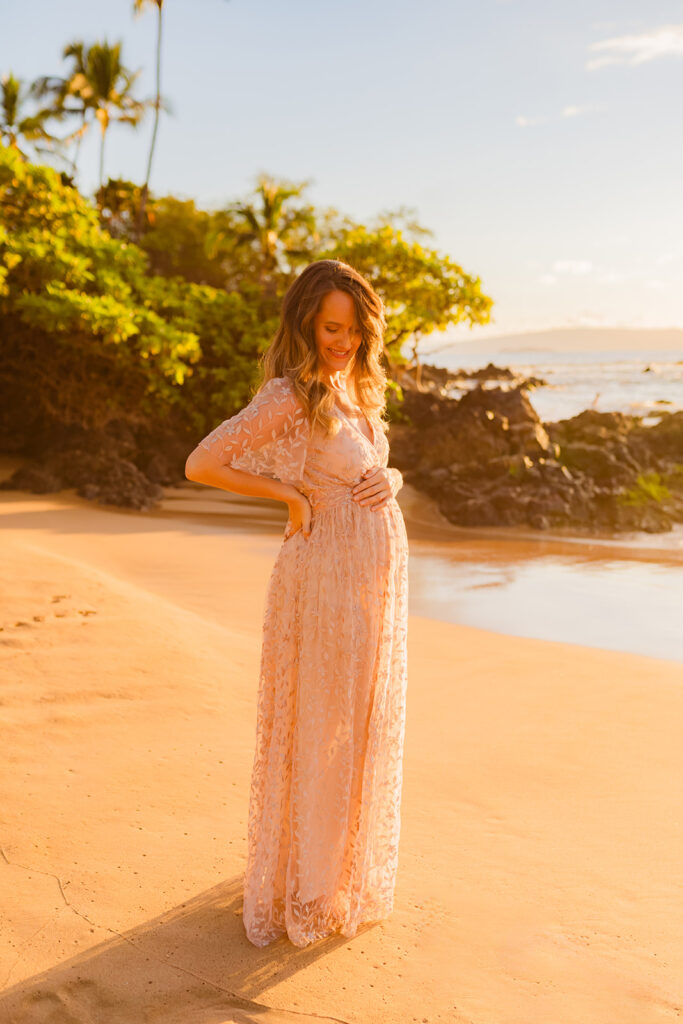 Woman wearing a beautiful champagne maxi maternity gown smiles down at pregnant belly on Maui