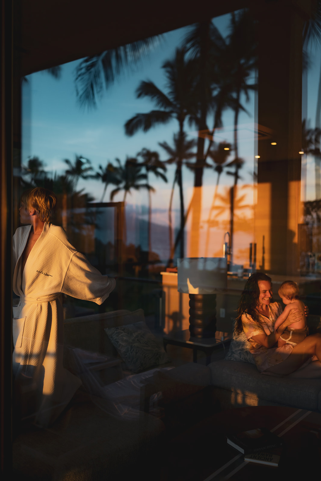 artistic reflection shot of mom holding and laughing with baby while her son plays in the foreground and palm tree reflections are shown on the window
