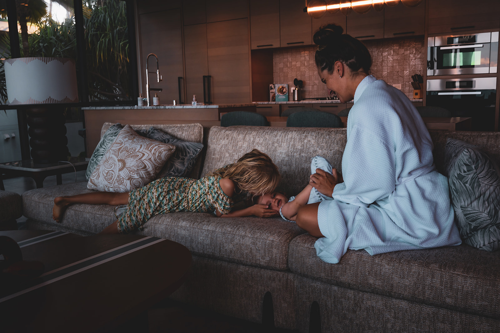 mother wears a white bathrobe and smiles at her daughter kissing baby's forehead