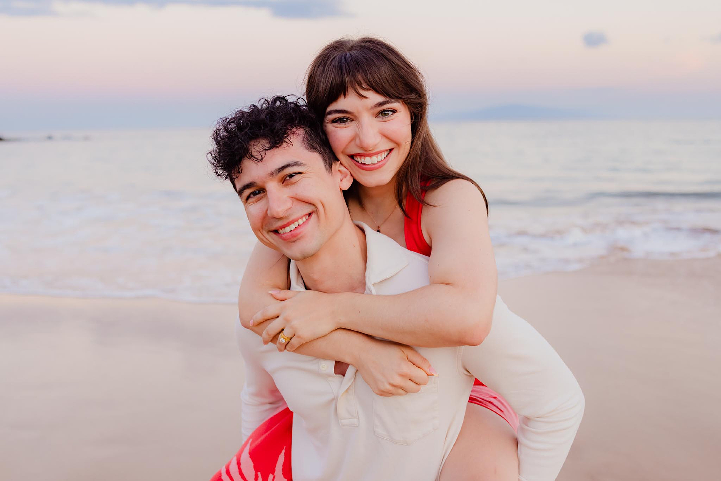 woman in red dress with bangs jumps on the back of her husband as they both smile on the camera